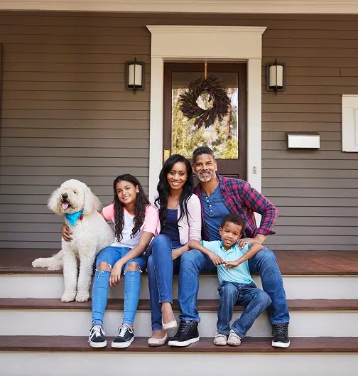 Family of four with white dog posing on front porch steps, brown horizontal siding house with cream trim, wreath on door